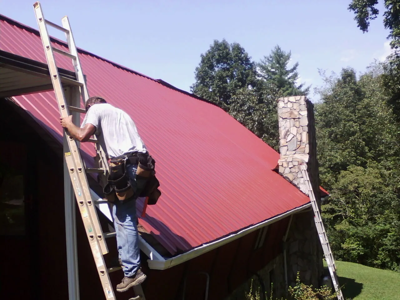 Roofer on a ladder beside a red metal roof and stone chimney — Soffit & Fascia Repair by Roofing Brothers in Grouse Creek